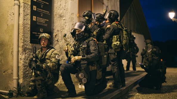 6 opérateurs AGIGN avec un militaire du dispositif Sentinelle, en extérieur. Ils échangent car un terroriste est retranché non loin d'où ils sont positionnés.