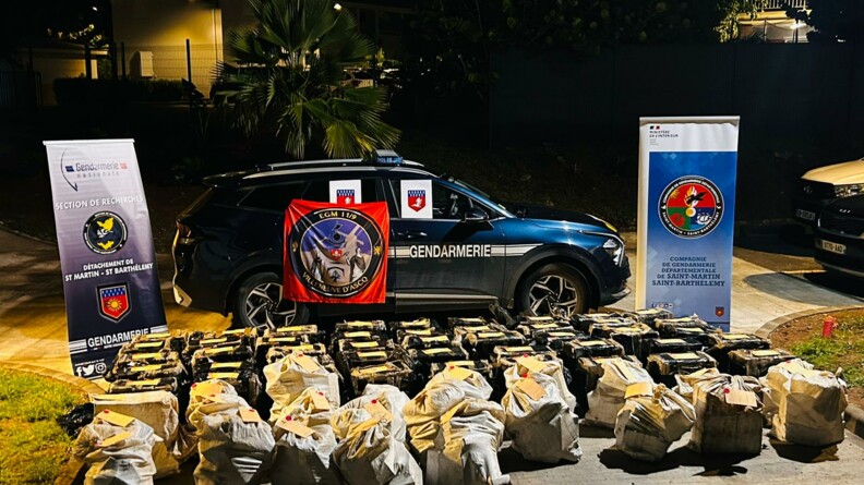 Photo de nuit montrant une voiture de gendarmerie devant laquelle plusieurs dizaines de sacs beiges sont posés. De chaque côtés, deux kakemono, un de la S.R de Saint-Martin ( à gauche) et un du COMGENDSBSM, sont positionnés.