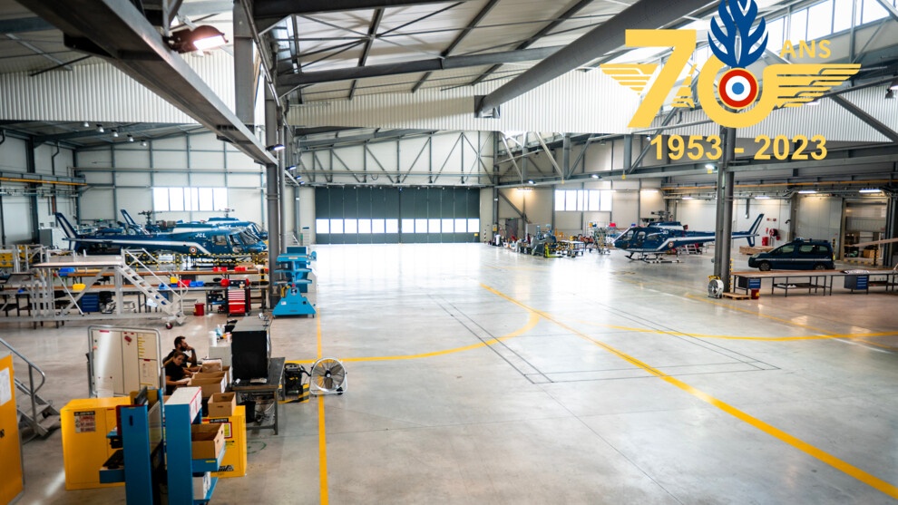 Photo présentant le hangar du CNaMAG accueillant les hélicoptères de la gendarmerie en maintenance. Les portes du hangar sont visibles au fond. Sur la gauche de la photo se trouvent deux appareils, ainsi que des militaires travaillant sur des postes de travail. À droite se trouve un hélicoptère ainsi qu'un véhicule gendarmerie.