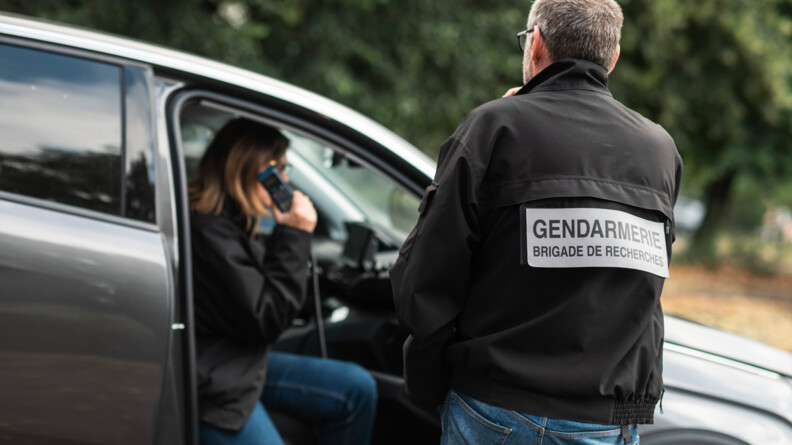 De dos un gendarme en civil, au blouson noir siglé "Gendarmerie brigade de recherches". Il regarde vers une gendarme dans la même tenue, dans un véhicule gris qui écoute la radio