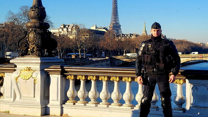 Un gendarme mobile debout devant la balustrade d'un pont de Paris. En arrière-plan, la Seine, Paris et la Tour Eiffel