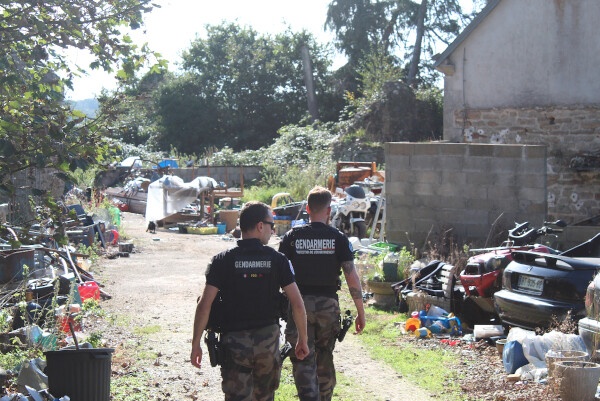 Deux gendarmes, vus de dos, se dirigent vers une habitation dont le terrain est jonché de déchets en tous genres et autres carcasses de véhicules.