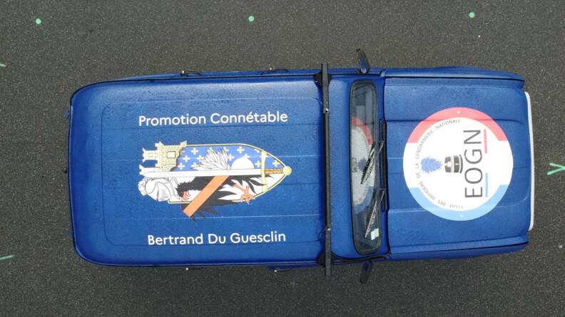 On aperçoit vue d'en haut une Renaud 4L bleu marine, couverte d'une grande enseigne sur le toit avec la mention "Promotion Connétable Bertrand Du Guesclin", et une seconde sur le capot portant la mention "Ecole des officiers de la gendarmerie nationale EOGN"