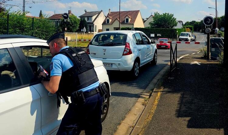 Un gendarme, vu de dos, dispense des conseils de prévention par la vitre du passager, à un conducteur arrêté derrière un autre véhicule, devant un passage à niveau (en arrière-plan), dont la barrière est baissée.