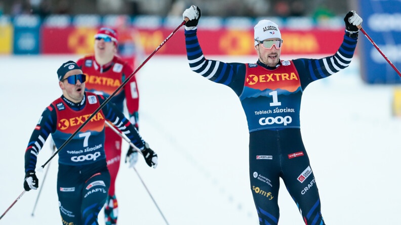 Sur la droite un skieur levant sees bras en signe de victoire. Au second plan à gauche, deux autres skieurs.