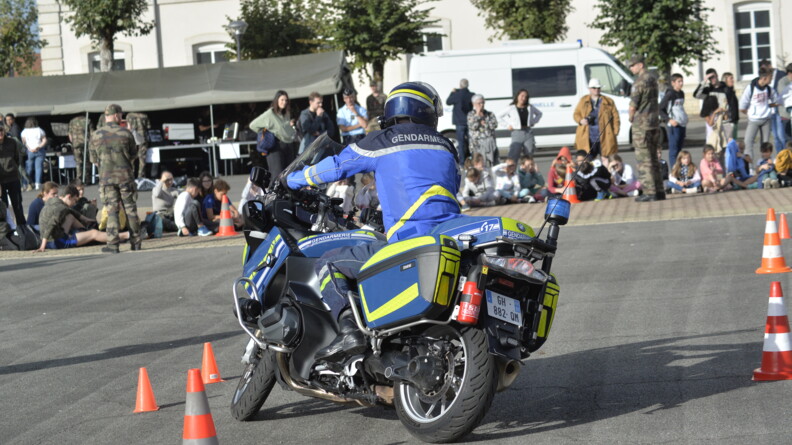 Un motard de la gendarmerie fait une démonstration sur sa moto en passant entre des plots, devant des spectateurs se tenant debout dans une cour.