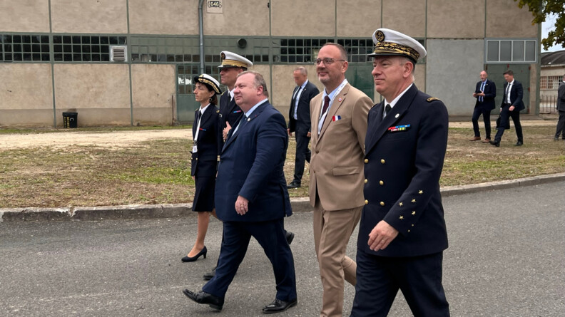 A l'extérieur, trois militaires avancent - deux hommes et une femme - entourantdeux hommes en tenue cicile ; l'un vêtu d'un costume bleu marine, l'autre vêtu d'un costume beige. En arrière plan, on aperçoit trois personnes qui marchent à l'écart du premier groupe.