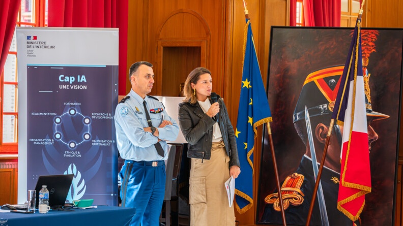 Une femme debout, aux côtés d'un gendarme, s'exprime avec un micro à la main. derrière elle, se trouvent un drapeau français et un drapeau européen