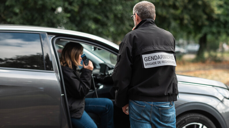 Un gendarme de dos en civil portant une veste noire siglée "Gendarmerie brigade de recherches" et jean, devant un véhicule gris avec sa collègue dans la même tenue