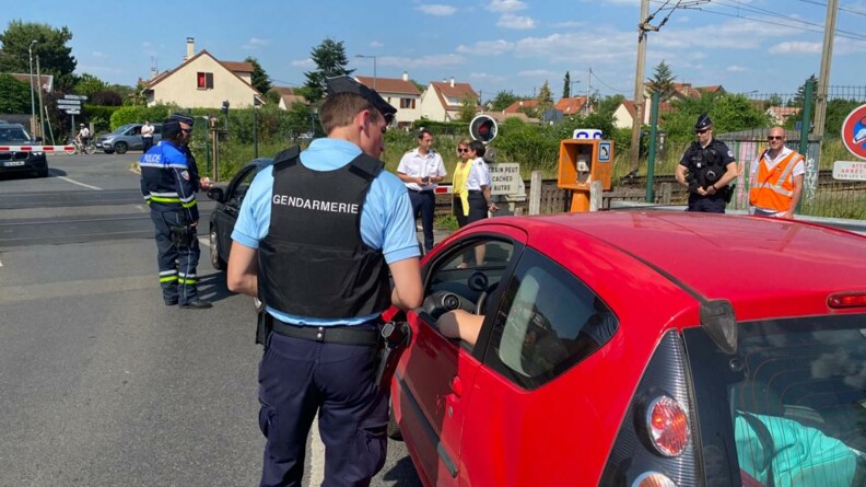 Un gendarme à côté d'un véhicule rouge à proximité d'un passage à niveau, rappelle au conducteur les règles de bonne conduite. En arrière-plan, d'autres gendarmes et des agents SNCF.