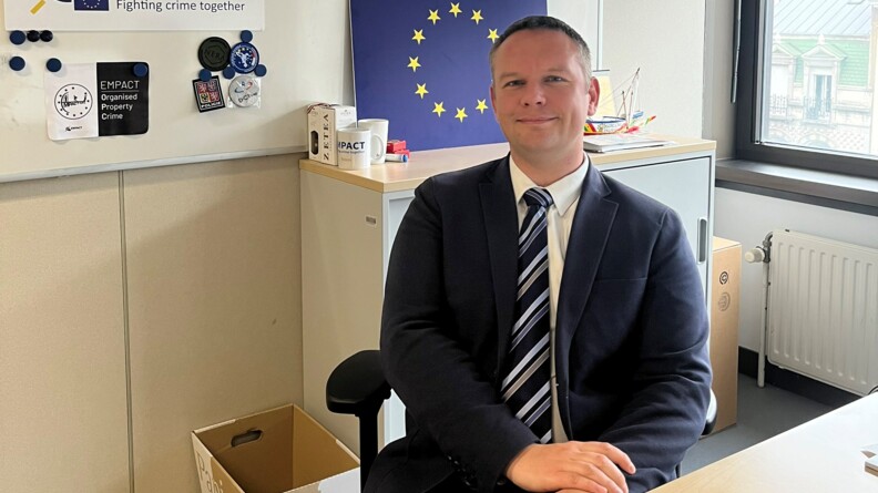 Portrait du CEN Simon carré, en costume bleu marine, chemise blanche et cravate rayée, assis derrière son bureau. Derrière lui, sur le mur, le drapeau européen et le logo EMPACT.