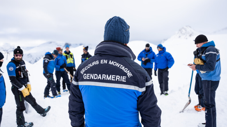 Sur un sol enneigné, un groupe d'hommes vêtus d'une tenue de ski de couleur bleue, se tiennent debout en cercle. Derrière eux se dressent les sommets enneigés.  Au dos de l'anorak de l'un d'eux, qui se tient au premier plan de la photo, on lit : Secours en montagne Gendarmerie