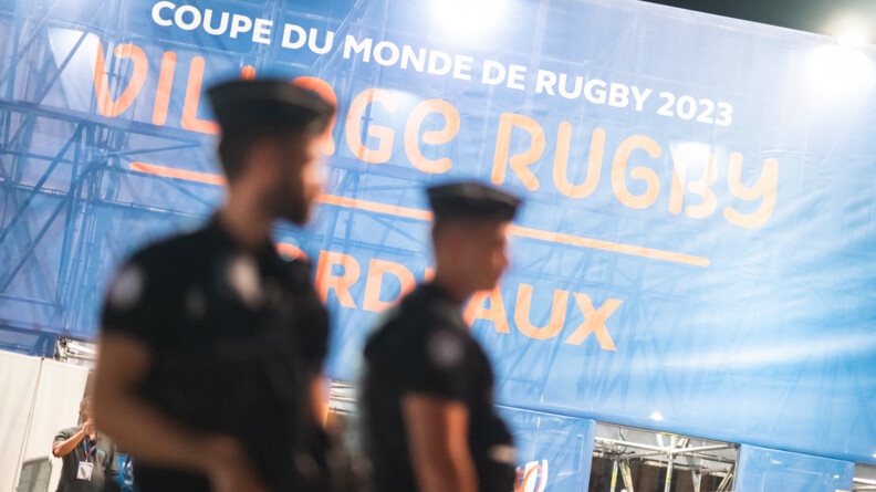 Au premier plan, deux gendarmes mobiles vus de profil. La netteté est faite sur l'arrière plan montrant la banderole d'entrée du village rugby.
