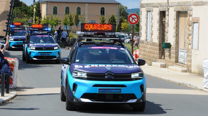Trois véhicules de la caravane du Tour de France féminin roulent dans un village