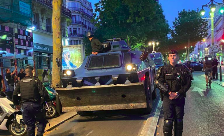 Deux gendarmes mobiles orientent la progression d'un véhicule blindé en plein coeur de la ville. Un troisième gendarme apparait au niveau de la tourelle.
