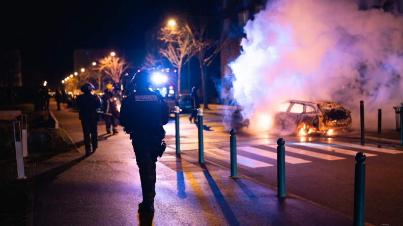 Dans la nuit, photo d'un gendarme mobile dans la rue. En fond, une voiture est entrain de bruler.