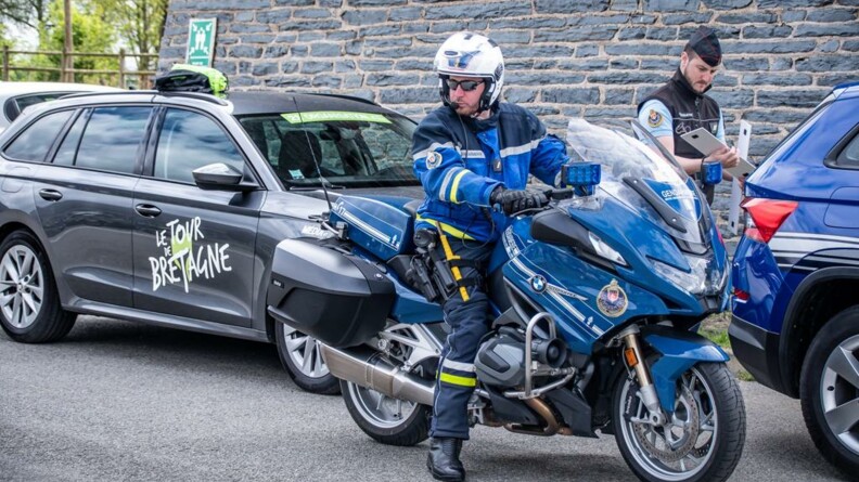 Motocycliste de la garde sur sa moto, pied à terre, surveillant la route. Derrière lui, sont garés un véhicule de l'organisation et un véhicule gendarmerie, à côté duquel un gendarme s'affaire.
