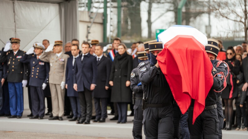 Au premier plan,  le cercueil du major Arnaud Blanc, recouvert du drapeau tricolore, est porté par six de ses frères d'arme du GIGN. Au second plan, les autorités civiles et militaires, au premier rang desquelles le président de la République, le ministre de l'Intérieur et des Outre-Mer, le ministre des Armées et le directeur général de la gendarmerie nationale.