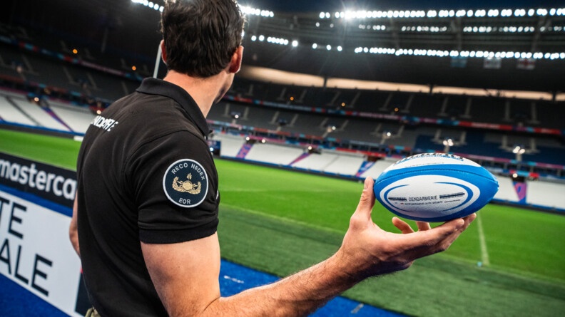 Gendarme EOR (Explosive ordonance reconnaissance) de profil tenant de la main droite un ballon de rugby aux couleurs de la gendarmerie devant la pelouse du Stade de France.