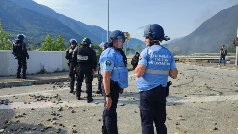 Deux gendarmes de l'équipe de liaison information sont sur une route jonchée de projectiles. Derrière eux, quatre gendarmes mobiles.