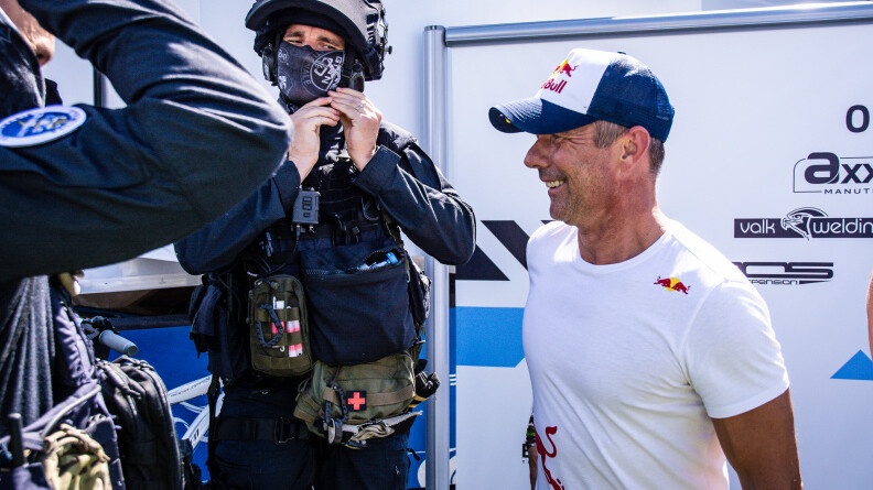 Un gendarme du GIGN en train de dégrafer son casque à la gauche de l'image. A sa droite, de profil droit, souriant, Sébastien Loeb en t-short blanc et casquette bleue et blanche red bull