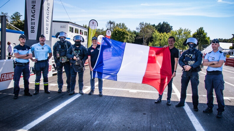 De gauche à droite, sur la piste de course : un gendarme départemental, un motard de la gendarmerie, deux militaires du GIGN, deux civils tenant un grand drapeau français, un troisime homme du GIGN, un dernier gendarme du GIGN