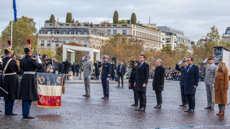Sur un sol pavé se tient debout le chef de l'Etat, Emmanuel Macron, entouré de personnalités officielles, civiles et militaires. Face à eux, se tiennent des hommes de la Garde républicaine, dont l'un d'eux tient un drapeau bleu blanc rouge. Derrière eux, on aperçoit des immeubles en pierre ainsi que des arbres feuillus.