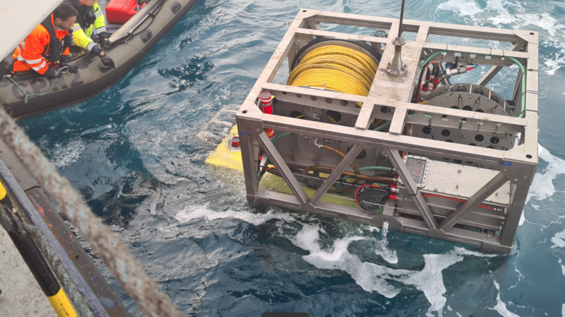 Une grosse caisse métallique, comportant un cable jaune, est extraite de la mer. Sur la gauche de l'image, deux hommes à bord d'un cannot pneumatique, supervisent la manoeuvre