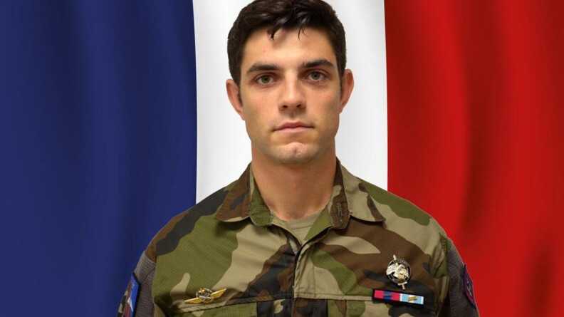 Un jeune militaire brun, yeux marrons, portant une tenue kakie devant un drapeau bleu blanc rouge recouvrant intégralement le fond de l'image.