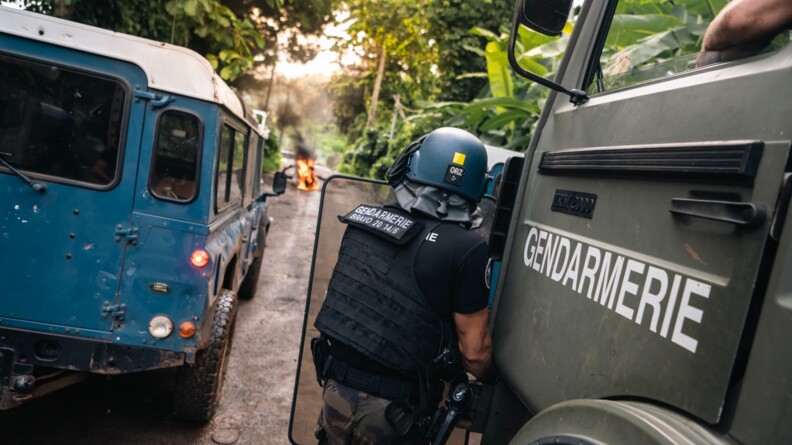 Un gendarme mobile vu de dos. Posté à côté d'un TRM kaki, il est derrière son bouclier. A sa gauche un Defender gendarmerie bleu. Dans le fond, une barricade en feu.