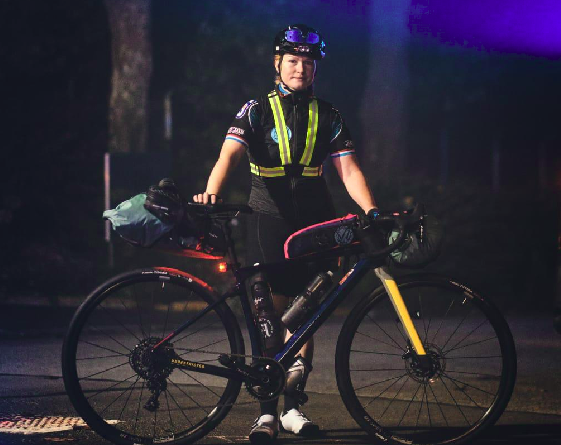 Une jeune femme cycliste pose de nuit devant son vélo. Elle se tient debout, une main posée sur la selle, l'autre sur le guidon de son vélo. Elle porte un casque et des lunettes par-dessus au niveau du front.