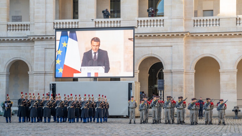 Un écran géant montre le Président de la république entrain de s'exprimer. En dessous, des gardes républicains et des militaires se tiennent au garde à vous.