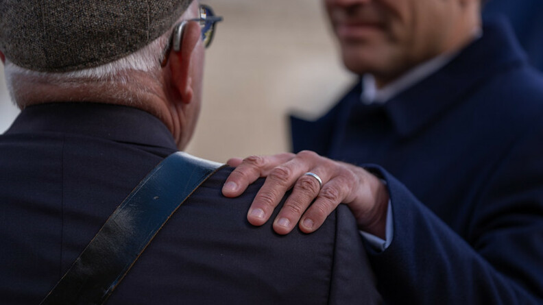 Une homme âgé apparait de dos, avec une veste bleue marine et un beret. Face à lui, se tient le président de la République, Emmanuel Macron, dont on aperçoit une partie du visage, et qui pose une main sur l'épaule gauche de l'homme.