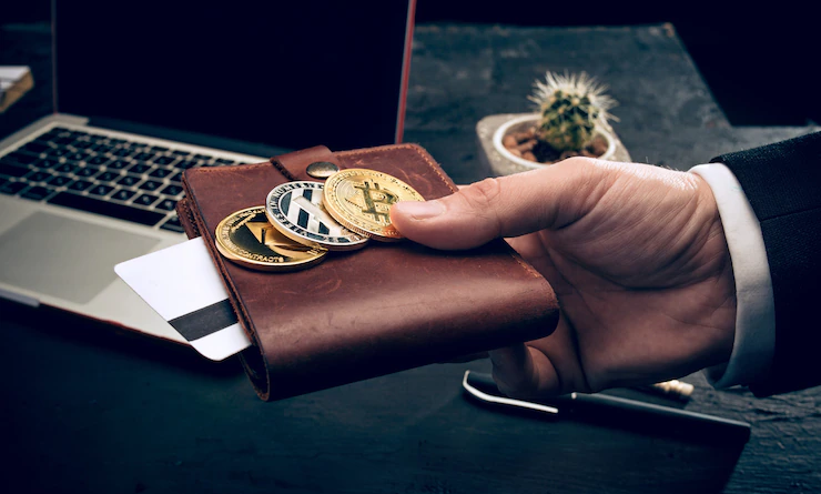 Photo d'illustration représentant un homme tendant un portefeuille avec des grandes pièces de monnaie, devant un bureau sur lequel est posé un ordinateur.