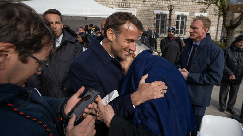 Le chef de l'Etat, Emmanuel Macron, tient dans ses bras, souriant, un vieil hmme recouvert d'une grande écharpe bleue, dissimulant son visage contre la poitrine du président.