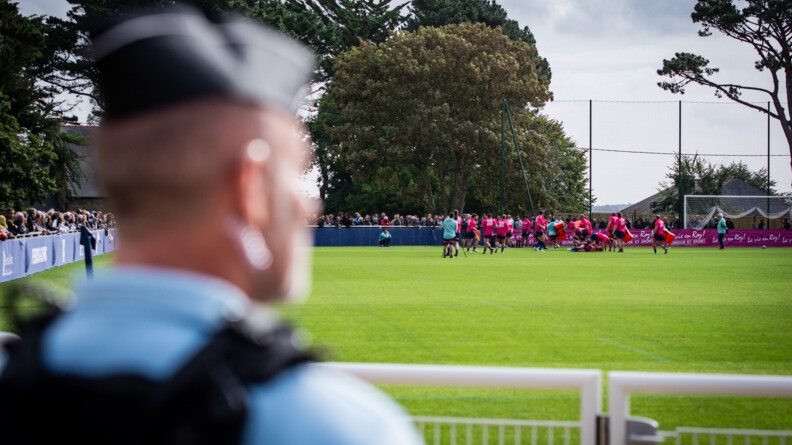 De dos en buste, flou, un gendarme, devant la pelouse où s'entraîne le XV du Chili. Autour du terrain, le public