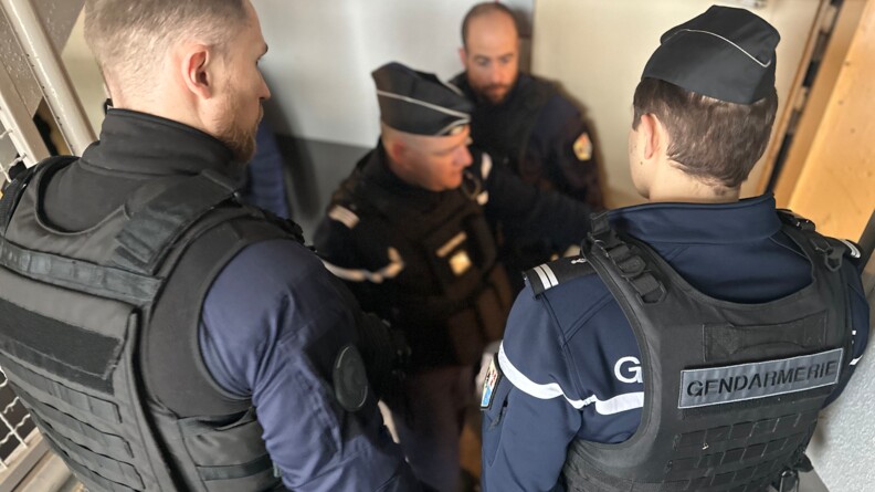 Plusieurs gendarmes en tenue se tiennent debout dans une cage d'escalier, au niveau d'une large porte de couleur beige entrouverte