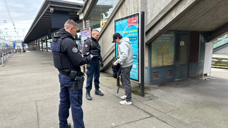 Deux gendarmes en train de contrôler un jeune homme vêtu d'un pantalon gris foncé, sweat gris clar et casquette noire dans une gare, sur les quais