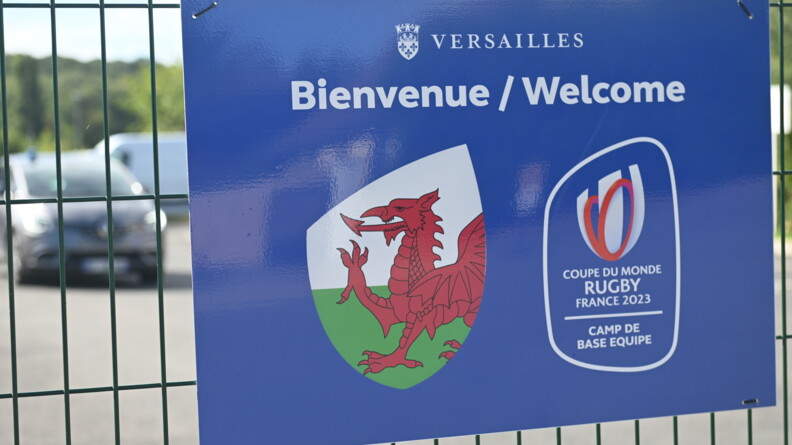 Panneau indiquant l'entrée du Camp de base de l'équipe du Pays de Galles à Versailles avec le logo de l'équipe (un dragon rouge sur fond vert et blanc) et celui de la Coupe du Monde de rugby France 2023, accroché sur une grille derrière laquelle on devine un véhicule de gendarmerie.
