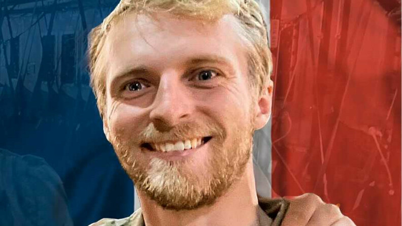 Photographie en couleur de Nicolas Mazier, jeune homme blond, barbu et souriant, posant en tenue militaire devant un drapeau français