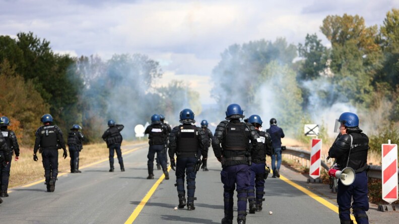Un groupe d'une douzaine de gendarmes mobiles vus de dos, avançant sur une route de campagne bordée d'arbres. Devant eux, un nuage de fumée blanche.