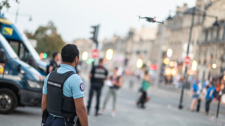 Au premier plan, un gendarme départemental vu de dos. Au second plan, dans le flou, un drone en vol et à gauche, l'avant de deux Irisbus de la gendarmerie mobile côte à côte et deux gendarmes mobiles.