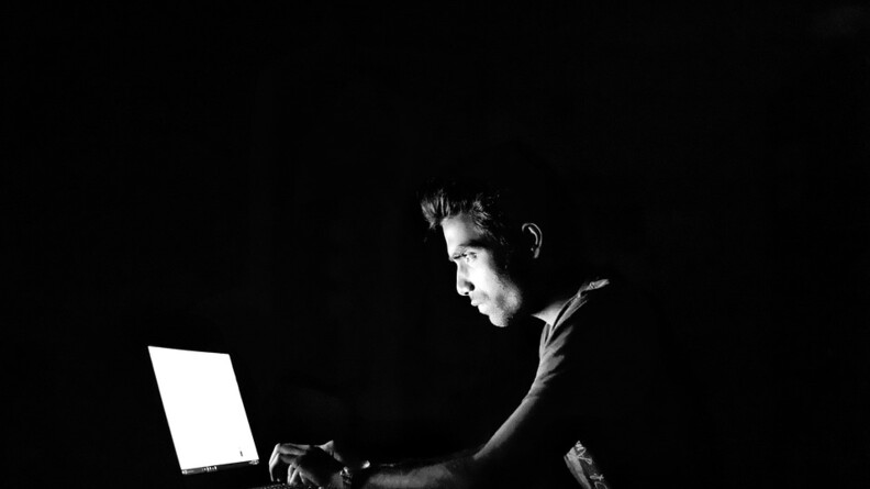 Image en noir et blanc montrant un individu piannotant sur un clavier d'ordinateur portable, le visage éclairé par l'écran de celui-ci.