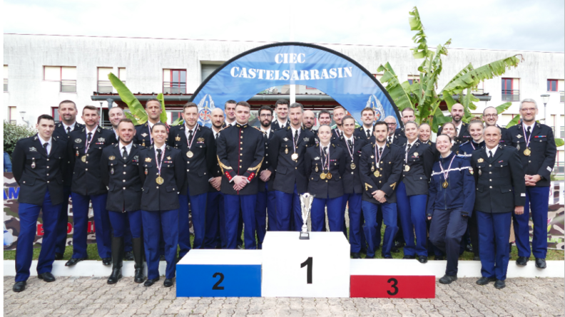L'équipe de gendarmerie du championnat de France militaire de badminton 2024, composée de 32 personnes, en uniforme, pose, souriante devant le podium revêtant les couleurs du drapeau français. Sur la premère marche est posée la coupe rempprtée par l'équipe. Derrière eux, une grande bannière bleue en arc porte la mention "CIEC Castelsarrasin"
