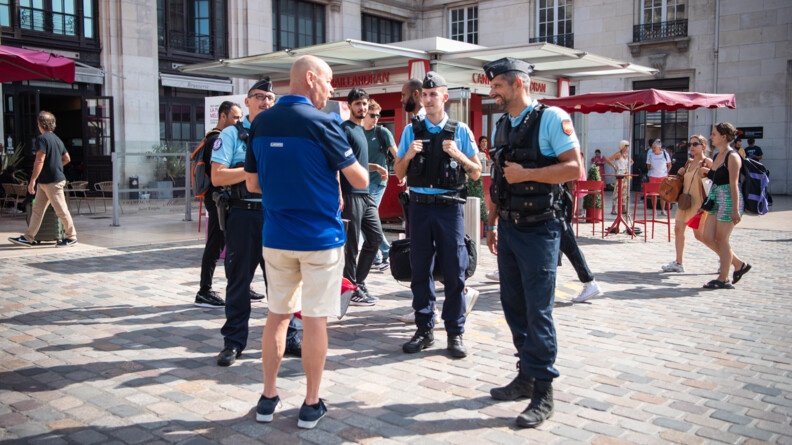 Trois gendarmes, vus de face, discutent avec un homme, vu de trois-quart dos, sur le parvis de la gare. En arrière plan, d'autres personnes passent.
