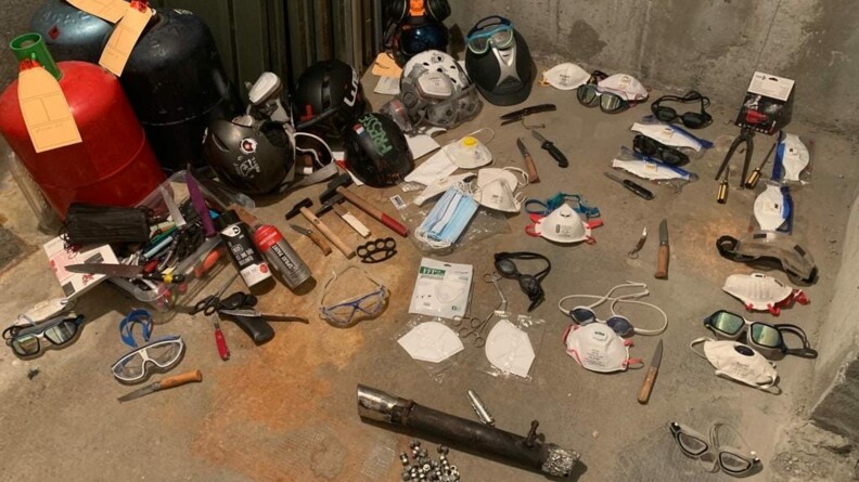Trois bouteilles de gaz, des casques et lunettes, des couteaux, marteaux, poings américains posés sur le sol d'un hangar.
