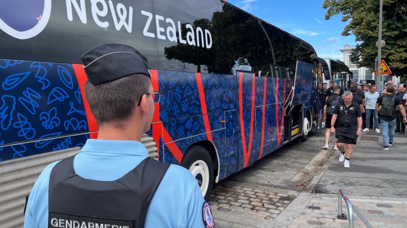 Au premier plan, un gendarme en tenue vu de dos. Devant lui, dans la longueur, un bus aux couleurs de la Coupe du Monde de rugby portant la mention "New Zealand". Dans le fond des joueurs et des membres du staff en train d'embarquer.