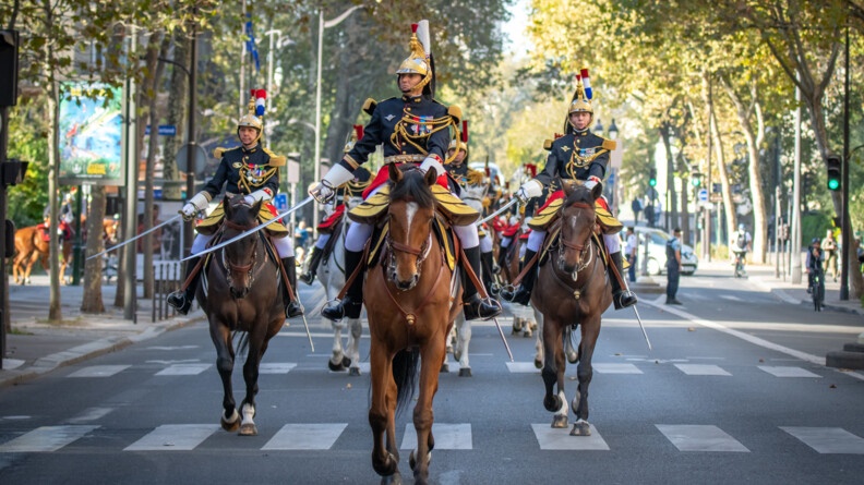 Vus de face, trois cavalier de la Garde républicaine en grande tenue, menant un peloton défilant dans les rues de Paris. Au centre, le colonel Leheup, chef du régiment de cavalerie de la Garde, et deux autres cavaliers qui saluent en baissant le sabre