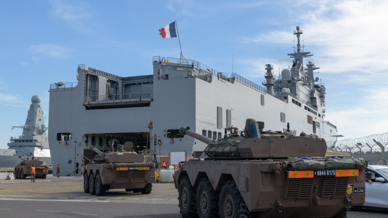 Des chars se dirigeant vers un bâtiment de la Marine gris surmonté d'un pavillon français