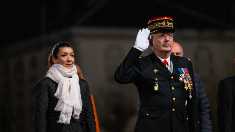 A gauche une femme en noir avec une écharpe blanche, les bras le long du corps, et le Directeur général de la gendarmerie nationale en vareuse, effectuant le salut militaire